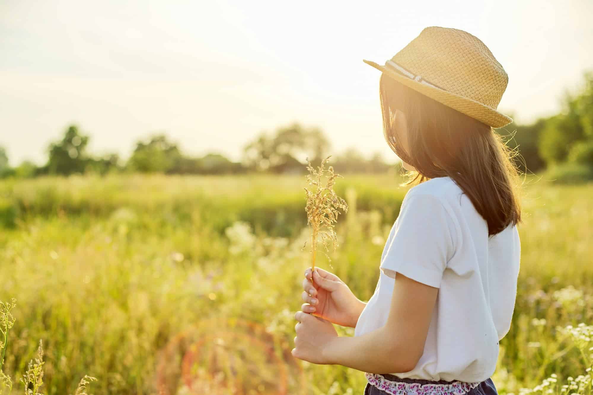 Summer nature, back view of child girl in straw hat tearing meadow grasses